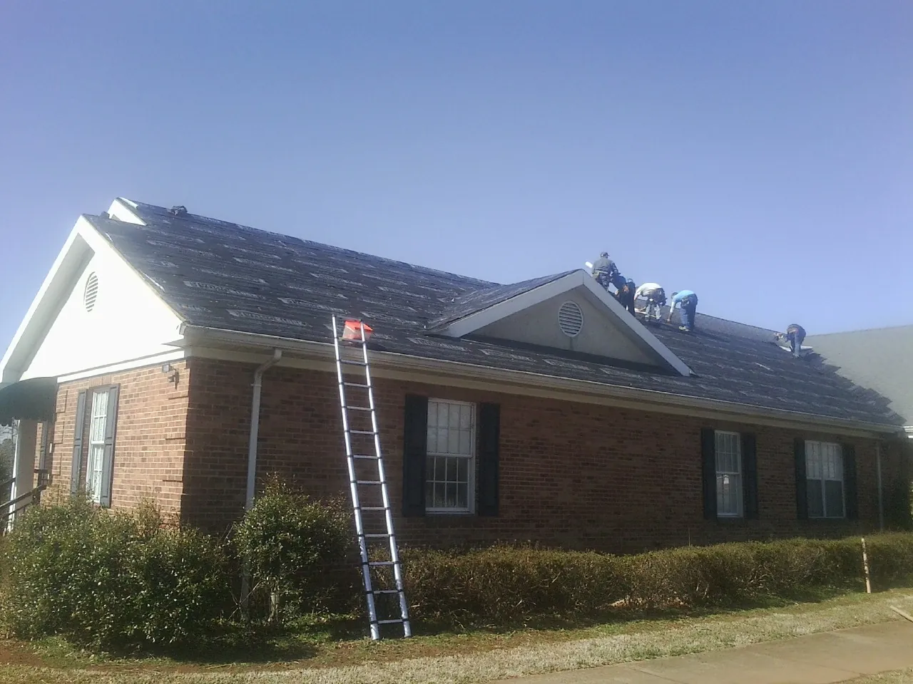 Crew tearing shingles off a brick ranch roof with a ladder set — Emergency Roof Repair work by our crew in Melba