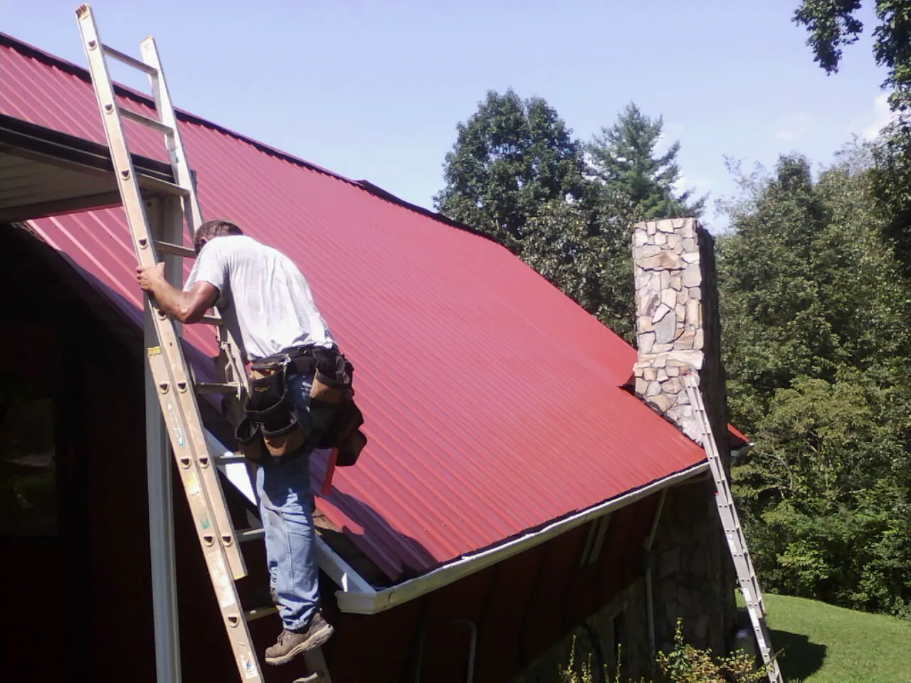 Roofer on a ladder beside a red metal roof and stone chimney — Soffit & Fascia Repair by Roofing Brothers in Melba